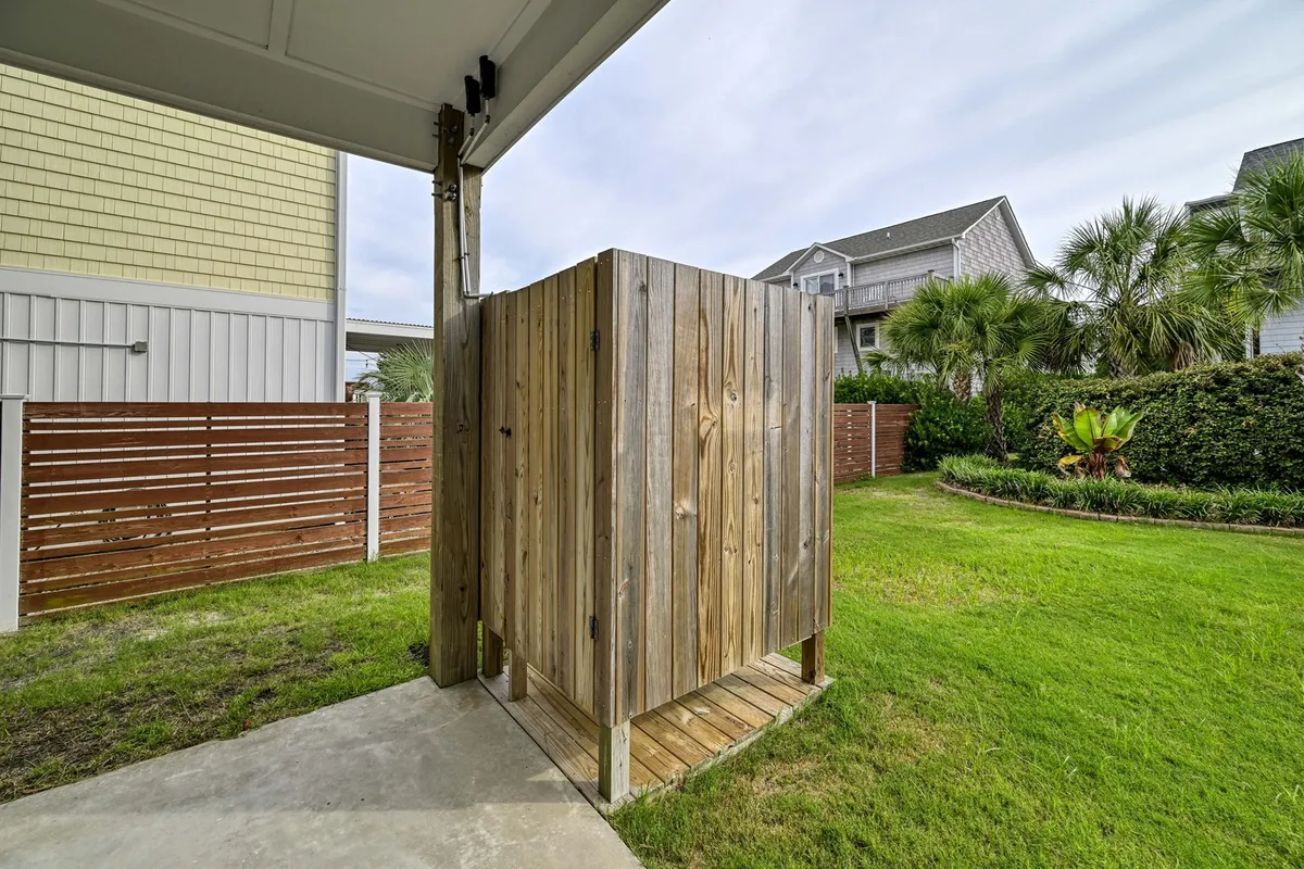 A private outdoor shower nestled against a fence, surrounded by greenery. The space looks refreshing and ideal for rinsing off after a day at the beach, embodying a blend of practicality and relaxation.