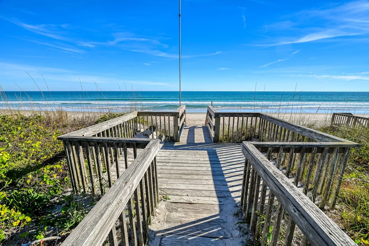 A wide view of a serene beach scene featuring a wooden walkway leading over dune grass toward the ocean, under a bright blue sky. The area is peaceful, with gentle waves lapping at the shore and plenty of space for families and pets to enjoy. Families can be seen building sandcastles and collecting seashells along the pristine shoreline.