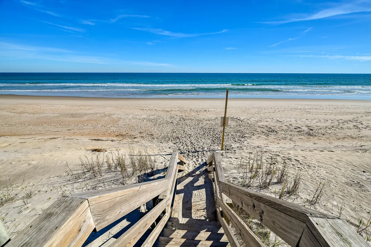 A classic exterior beach access scene showing wooden stairs leading down to the sandy shore, with gentle ocean waves and a bright blue sky in the background. The pathway invites visitors and their pets to start their day with a walk to the ocean.