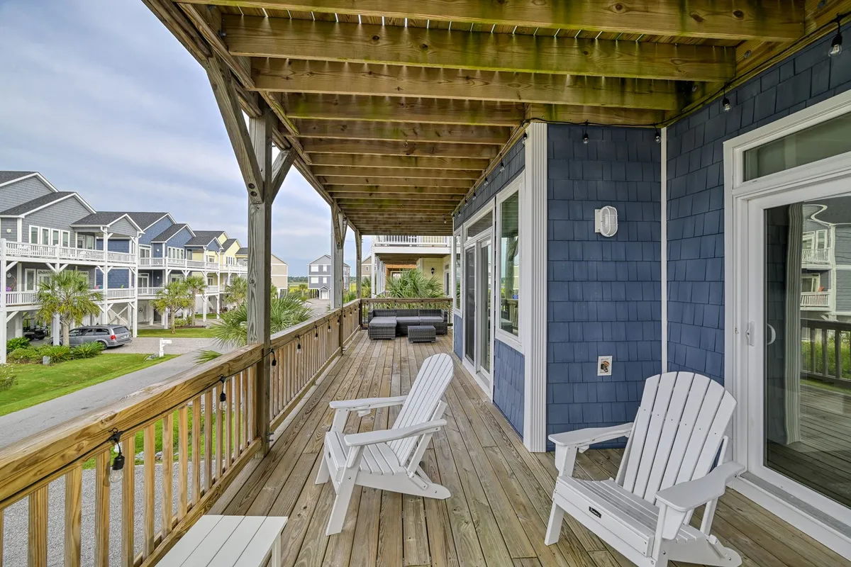 A large wooden deck with white rocking chairs, surrounded by greenery and neighboring houses, offering a tranquil space for relaxing, entertaining, or enjoying views. The porch feels like an extension of the indoor social spaces, ideal for letting the salty air flow in.