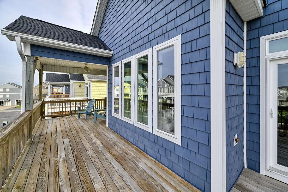 This exterior shot features a blue shingle house with a spacious wooden deck furnished with Adirondack chairs. Large windows overlook the deck, inviting the outdoors in and signaling a space designed for relaxation and social gatherings. The mood is calm and welcoming, emphasizing the connection between inside comforts and open-air enjoyment.