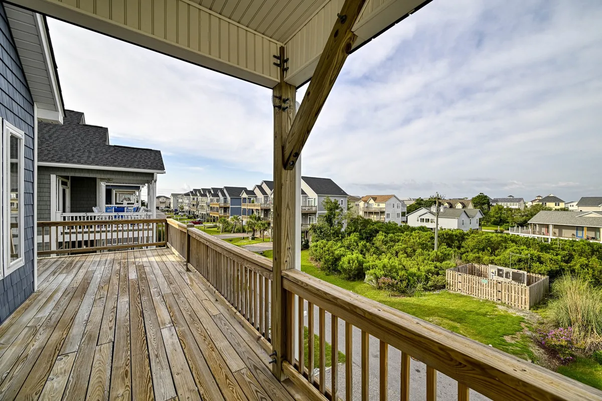 This image shows a wooden deck with railings, surrounded by greenery and neighboring houses. The space is ideal for morning coffee, hosting small gatherings, or relaxing in the sun. The setup suggests a welcoming atmosphere for outdoor group activities and enjoying the local environment.