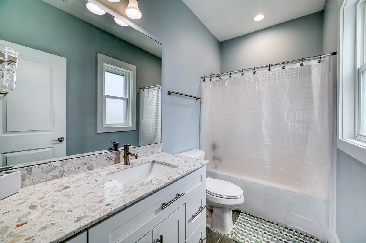 A stylish bathroom featuring a large mirror, granite countertop, inviting bathtub, and a window allowing natural light to enhance the space. The blend of modern fixtures and tranquil tones sets the scene for ultimate relaxation.