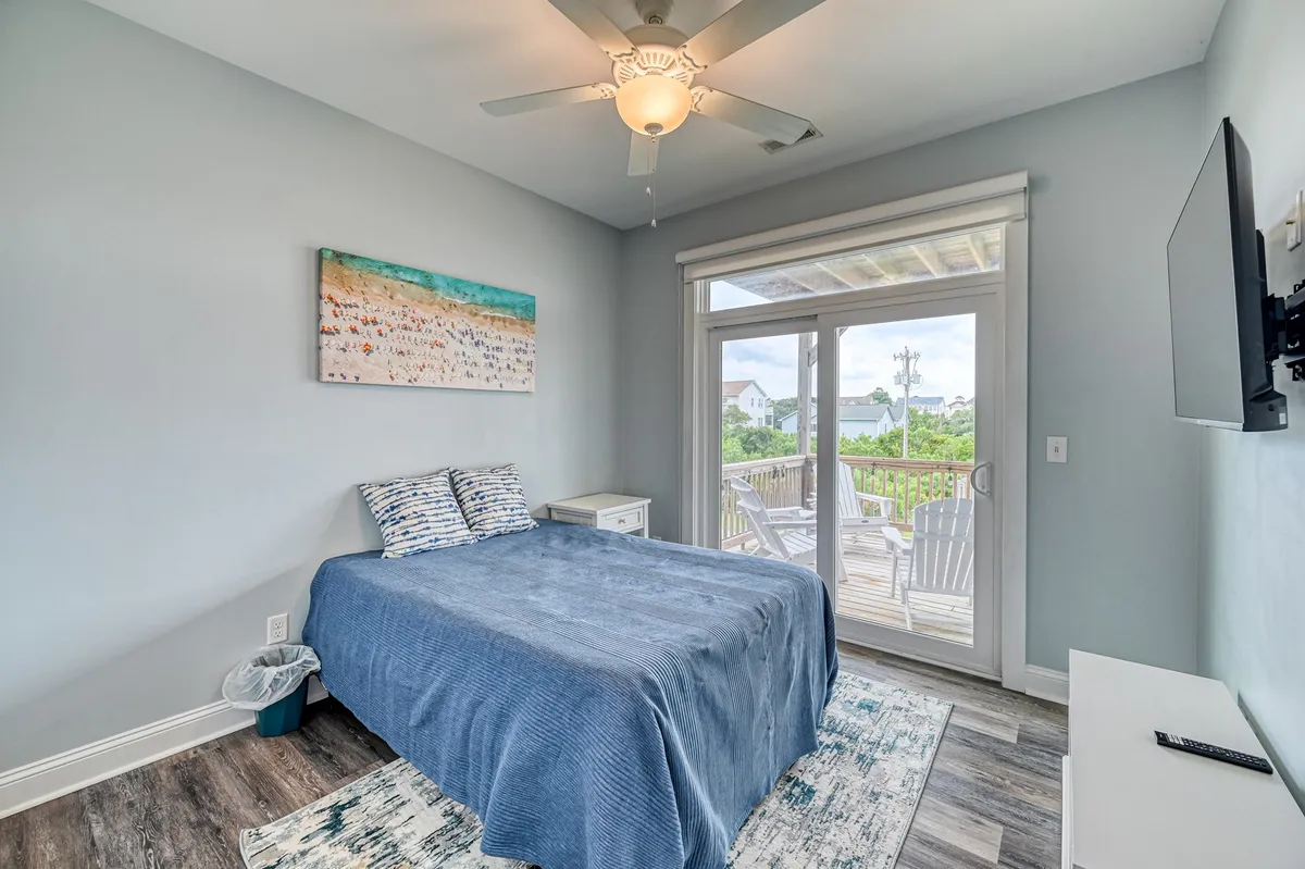 A tranquil bedroom featuring a comfortable bed, sliding glass door leading to an outdoor deck, and beach-themed artwork. The room is bathed in natural light, inviting guests to sit with a morning coffee or step outside to enjoy the fresh air and ocean views.