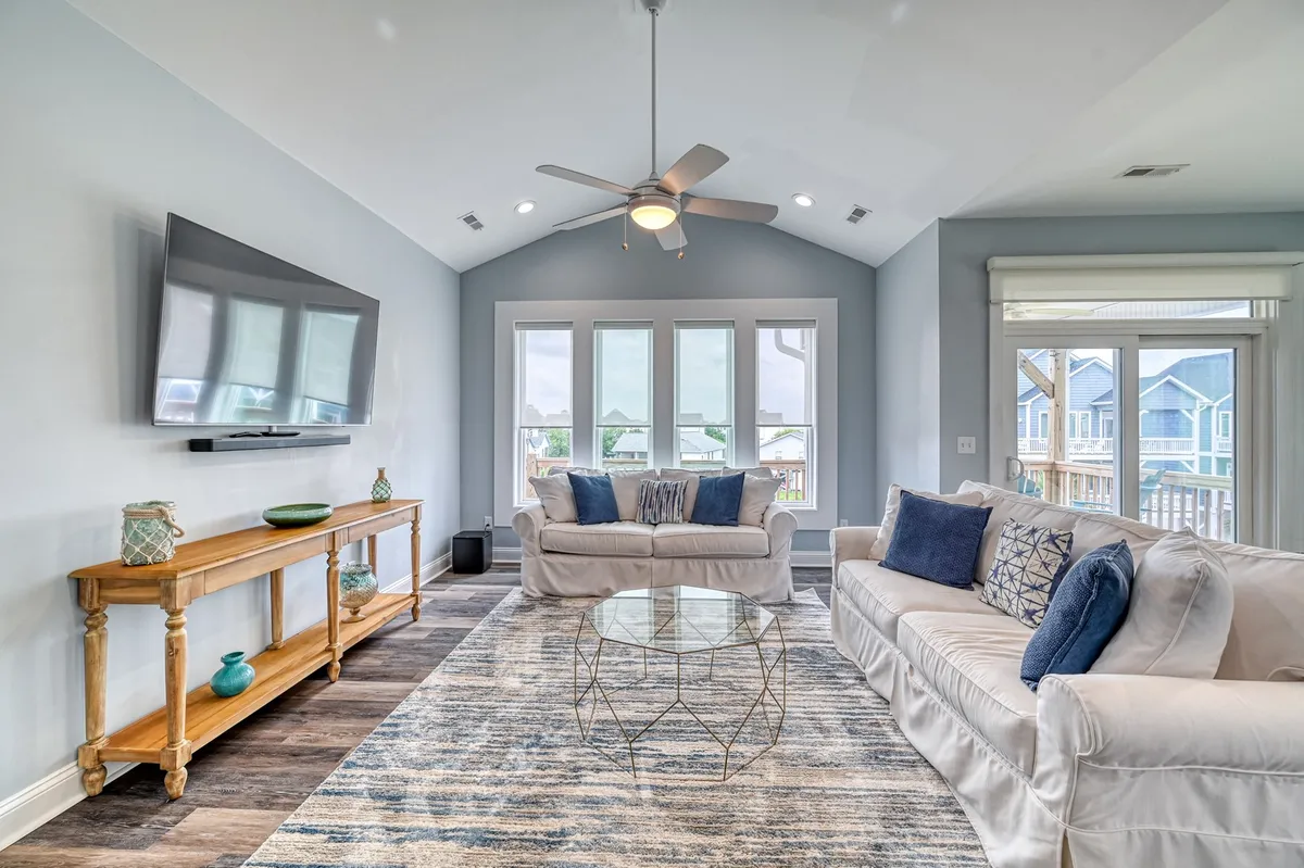 The image features a sunlit living room with a plush blue-cushioned sofa, a modern geometric coffee table, a wall-mounted television, and expansive windows that flood the space with natural light—hinting at sunrise views. The setup invites guests to unwind, socialize, or enjoy cozy entertainment in a comfortable setting.