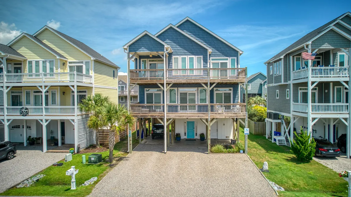 This image shows a cheerful exterior scene with colorful houses lined up, palm trees, and a spacious gravel driveway. The driveways accommodate multiple vehicles, and there are porches where people are seen relaxing and socializing.