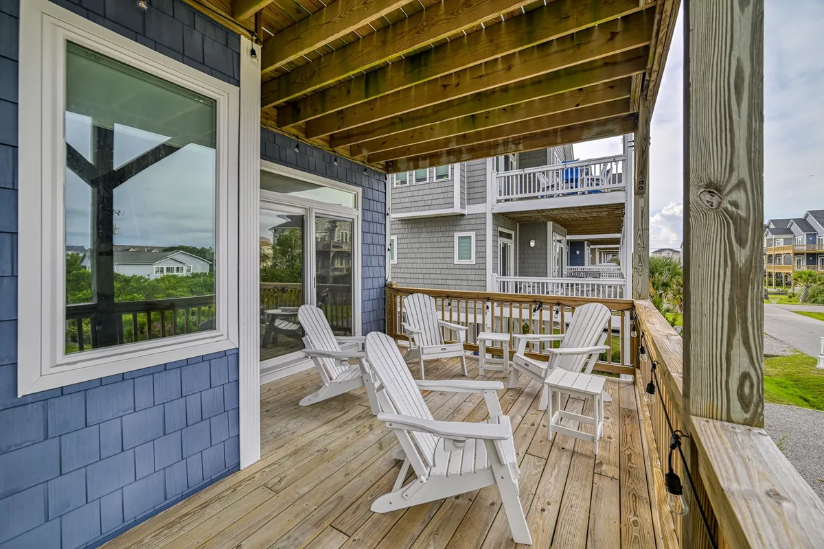 A wooden deck is set against a backdrop of greenery and neighboring houses, with several white Adirondack chairs arranged for lounging. The scene feels relaxed and welcoming, perfect for morning coffee, quiet reading, or evening gatherings, and it emphasizes the versatility of the porch area.