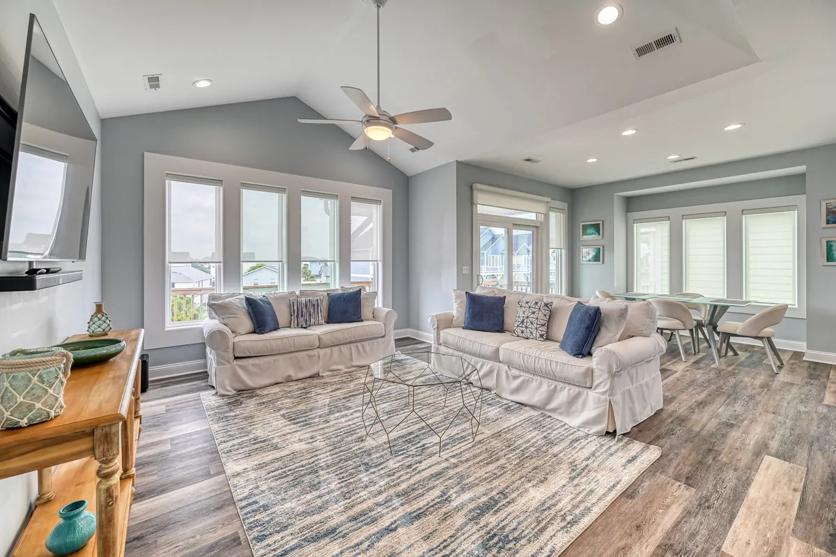 A welcoming living room with plush sofas arranged around a coffee table, a television, and large windows allowing natural light to flood the space. The layout encourages relaxation and group activities, like movie nights or board games, while the furnishings have a coastal, comfortable vibe.