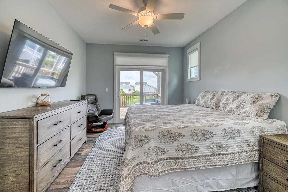 A serene bedroom featuring a large bed with decorative bedding, a sizable window filling the room with natural light, a wall-mounted television, and a comfortable chair positioned for reading or relaxing. The soothing colors and inviting textures make it an ideal sanctuary for rest.