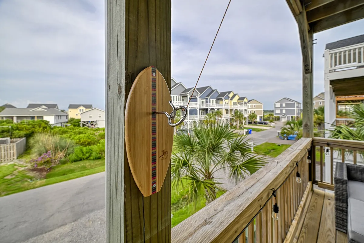 A sunny porch scene with a wooden pulley, surrounded by palm trees and neighboring houses. The porch area is casual and inviting, perfect for relaxing after a day at the beach, watching sunsets, and socializing with family or friends. The neighborhood looks peaceful and safe, ideal for families seeking tranquility and community.